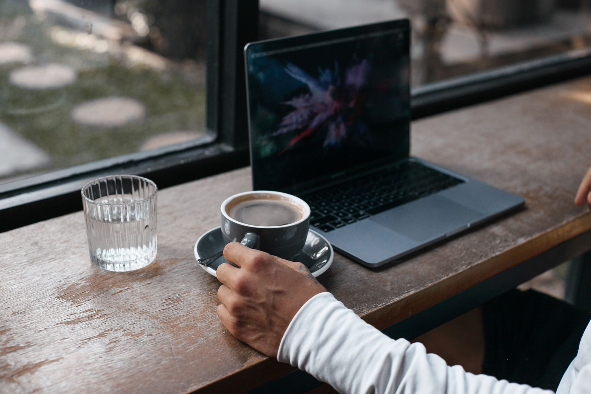 Mano sosteniendo una taza de café junto a un portátil sobre una mesa de madera junto a una ventana.