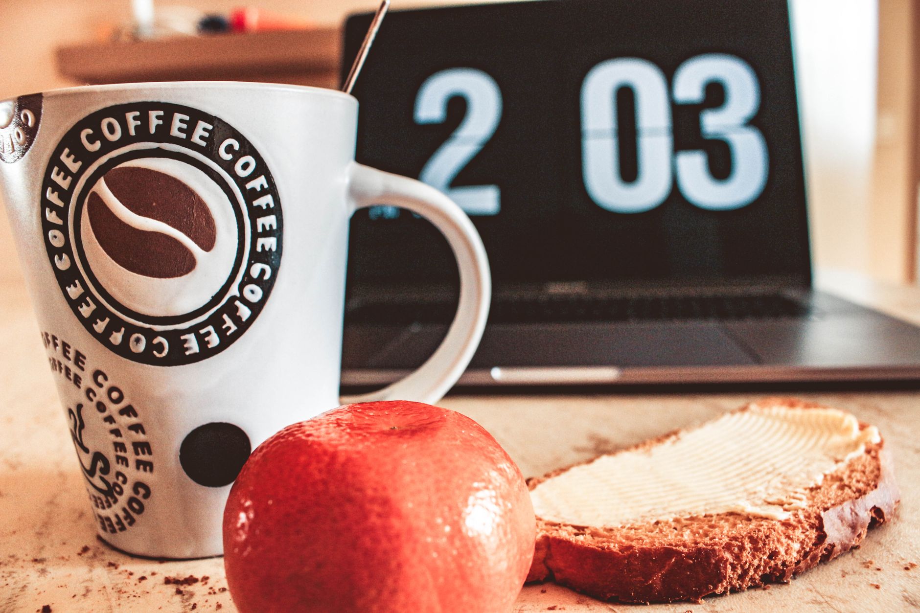 Mesa de desayuno acogedora con café, pan y fruta junto a un portátil.